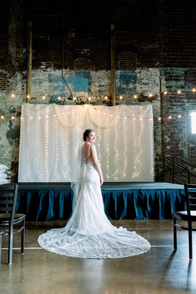 Photo of the bride in her wedding dress in front of the stage where she would be married. Party lights hang from the ceiling, layers of paint from plays past on the exposed brick wall behind her.